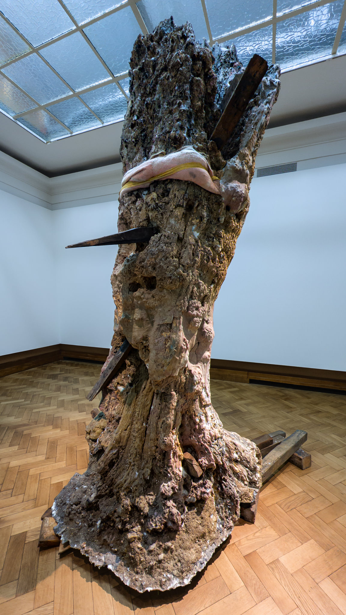 The image shows an artwork from the exhibit "Khorós: A Mesmerizing Journey Through Berlinde De Bruyckere's Vision". The piece features a large, textured, tree trunk-like structure standing upright in a bright, white-walled gallery room with a wooden floor and a glass ceiling.

The sculpture resembles an ancient, decayed, and weathered tree stump. It's organically shaped, with rough, jagged bark and surfaces that look almost fossilized. Several dark wooden beams pierce through the trunk at different angles, adding a harsh, almost violent contrast to the organic form. Wrapped around the upper part of the trunk is a soft, pinkish-beige fabric or textile, draping delicately, creating an eerie juxtaposition of tenderness and brutality. The base of the sculpture is rugged and irregular, as though it was unearthed directly from the ground.

The overall mood of the artwork feels raw, contemplative, and haunting. Berlinde De Bruyckere is known for exploring themes of vulnerability, decay, and the delicate balance between destruction and care, which is clearly reflected in this powerful piece.

Would you like a deeper interpretation of the symbolism or details about the materials used?