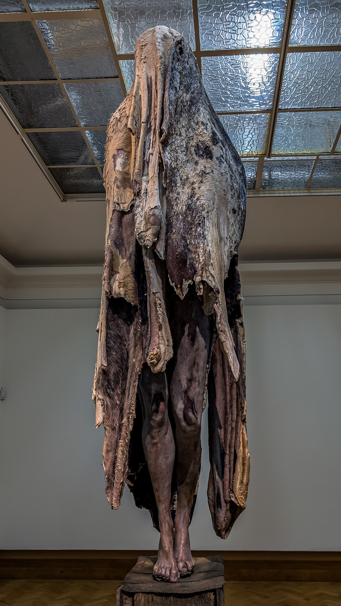 The sculpture depicts a haunting figure shrouded in a tattered, flowing covering that resembles skin or fabric, evoking a sense of decay and vulnerability. Standing upright, the figure has a distressed, almost ghostly appearance, with the cloak hanging loosely, draping down in uneven layers. The feet, bare and grounded on a rough wooden pedestal, contrast sharply with the texture of the covering. The expressionless face is obscured, adding to the unsettling nature of the piece. Set against the backdrop of a gallery with reflected light from a textured ceiling, the artwork invites contemplation on themes of identity, mortality, and the transient nature of existence.