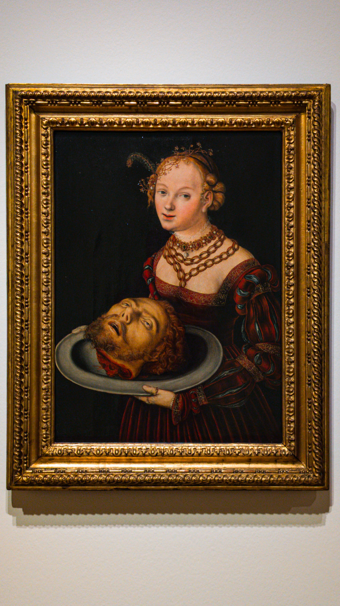 The painting features a young woman elegantly dressed in rich red and black attire, adorned with intricate jewelry around her neck and in her hair. Her expression is calm and composed as she gazes directly at the viewer, suggesting a mix of pride and defiance. In her hands, she holds a large, shallow platter containing a severed head with long hair and a beard, adding a dramatic and unsettling contrast to her serene demeanor. The dark background emphasizes the vivid colors of her clothing and the details of her accessories, creating a striking focal point on the unsettling subject of the head. The ornate gold frame enhances the opulence of the piece while framing the complex themes of beauty, tragedy, and power.