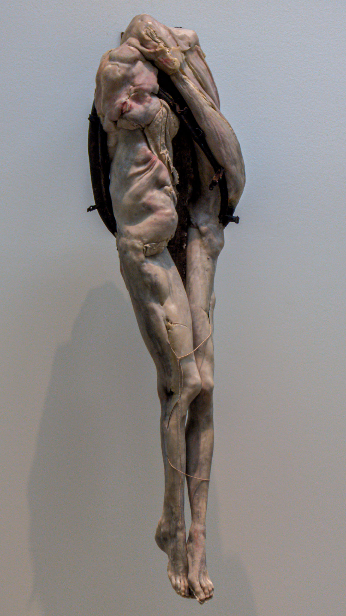 The sculpture presents a striking and unsettling figure, reminiscent of a human form yet infused with a sense of distortion and fragility. The body is elongated, with limbs that appear contorted and intertwined, conveying a tension between movement and stillness. The surface texture suggests flesh, complete with detailed features that evoke both realism and a hint of decay.

Ropes or straps seem to hold the figure in place, adding an element of restraint. The legs are thin and elongated, ending in small, bare feet that touch the ground. The overall presentation prompts reflection on themes of vulnerability, existence, and the human condition, creating an emotional resonance that invites deeper contemplation from viewers. The neutral backdrop emphasizes the figure's raw, visceral quality, allowing its haunting presence to dominate the space.